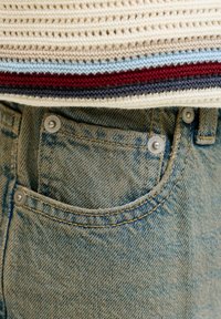 Denimfarkute lähedalt vaadatuna, mille taskul on metallist naelad, ning kootud kampsuni ääres, millel on punased, sinised ja kreemjad horisontaalsed triibud.