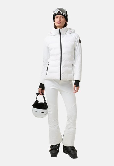 Biała ocieplana kurtka narciarska z czarnym zamkiem i detalami, w zestawie z białymi spodniami narciarskimi. Model trzyma czarno-biały kask, nosi gogle narciarskie.