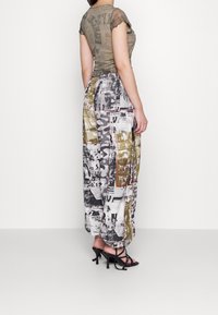 Przezroczysta oliwkowa bluzka z krótkimi, falbaniastymi rękawami; wzorzyste, szerokie spodnie w kolorze czarnym, białym i złotym o luźnym kroju; czarne sandały na obcasie z paskami.