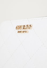 Portefeuille en matériau synthétique matelassé blanc avec le logo doré "GUESS". Présente un motif en diamant et des coutures assorties. Texture lisse.