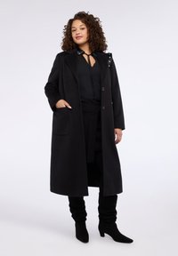 Czarny płaszcz trencz z wełny o dopasowanym kroju, dużym kołnierzem i kieszeniami. Zestawiony z czarnymi botkami i bluzką z wiązaniem. Gładka faktura.