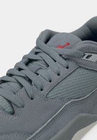 Gros plan d'une chaussure de sport grise avec des panneaux en tissu texturé, des lacets épais et un petit logo rouge brodé sur la languette.