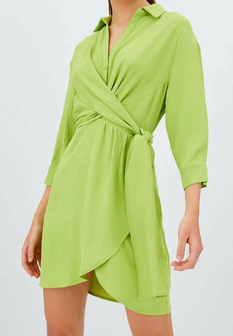 Abito avvolgente verde a maniche lunghe in tessuto leggero; presenta un colletto, una cintura in vita e un orlo asimmetrico. Tessuto liscio con un colore uniforme.