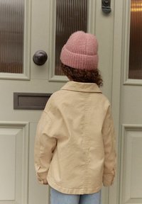 Chaqueta beige oversized con cuello y puños abotonados, complementada con un suave sombrero de punto rosa. Tela lisa contra una puerta texturizada.