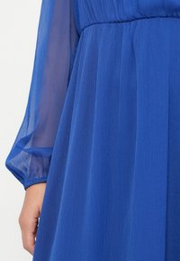 Close-up da mão de uma pessoa perto da cintura de um vestido azul com mangas longas transparentes e cintura franzida.