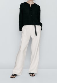 Cardigan nero a maglia con scollo rotondo e due tasche frontali, abbinato a pantaloni larghi a righe crema e sandali neri bassi.