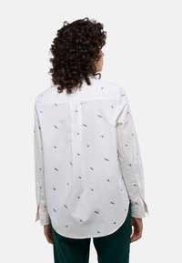 Chemise blanche à manches longues avec des motifs brodés verts. Col classique, poignets à boutons et ourlet arrondi conçu pour un ajustement décontracté.