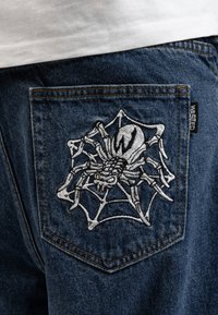 Jeans i denim har en detaljerad vit spindel broderad på bakfickan, kopplad till en spindelnätsdesign med svarta accenter och synlig sömnad.