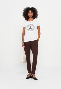 Mulher com cabelo encaracolado vestindo uma t-shirt branca com o logo de âncora, calças castanhas, sapatos de salto pretos e a segurar uma bolsa beje claro, contra um fundo branco.