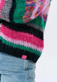 Barevný pletený svetr s pruhy v růžové, zelené, černé a šedé barvě, s malou růžovou štítkem na lemu. Textura působí měkce a útulně.