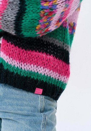 Kolorowy sweter na drutach z paskami w kolorach różowym, zielonym, czarnym i szarym, z małą różową metką przy krawędzi. Tekstura wygląda na miękką i przytulną.