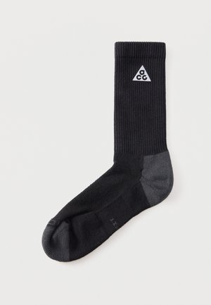Șosetă sportivă neagră cu călcâi și vârf gri, cu un logo alb ACG sub formă de triunghi lângă manșeta superioară pe un fundal simplu deschis.