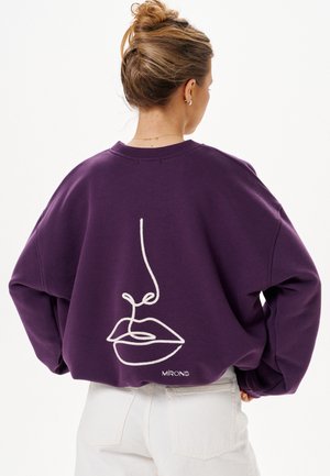 Donna con capelli raccolti in uno chignon indossa una felpa viola con un disegno a linea continua bianca di labbra e naso sulla schiena.