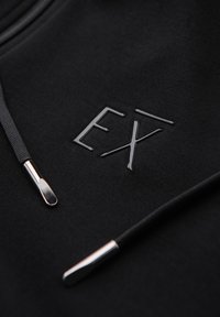Tissu de sweat à capuche noir montrant un logo "EX" brillant et des cordons noirs avec des embouts métalliques argentés en gros plan.