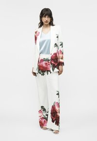 Mujer vistiendo un traje blanco con grandes flores rosas y rojas, blusa azul claro y tacones abiertos blancos, posando contra un fondo liso.