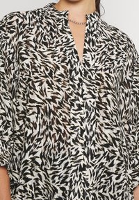 Schwarz-weiße gemusterte Bluse mit Stehkragen, kurzen Ärmeln und einer vorderen Knopfleiste. Leichtes, strukturiertes Gewebe mit abstraktem Design.