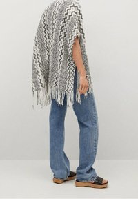 Dziany poncho w czarno-biały wzór w zygzaki, z frędzlami na brzegach. Połączone z luźnymi, jasnoniebieskimi jeansami i brązowymi drewnianymi sandałami na platformie.