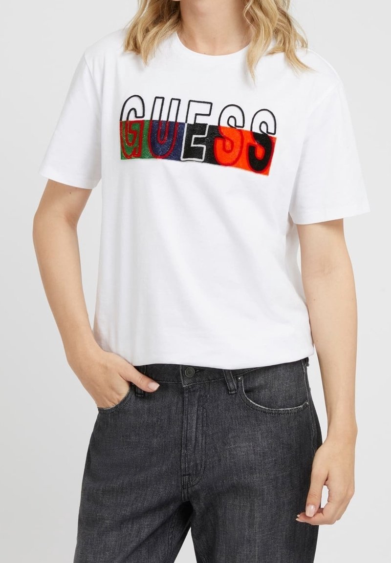 T-shirt en coton blanc avec un logo "GUESS" multicolore en lettres majuscules. Manches courtes, coupe décontractée, associé à un jean gris foncé.
