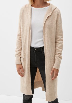 Femme portant un long cardigan à capuche beige par-dessus une chemise blanche et un jean noir, debout devant un fond blanc uni.