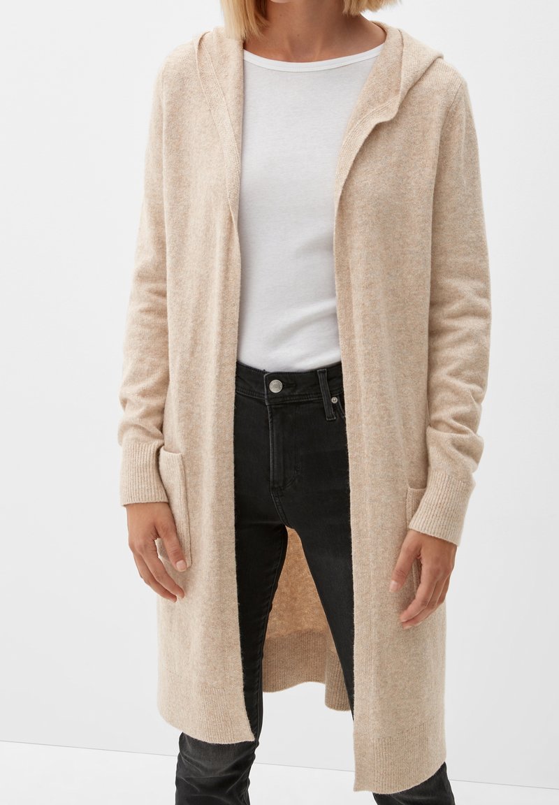 Femme portant un long cardigan à capuche beige par-dessus une chemise blanche et un jean noir, debout devant un fond blanc uni.