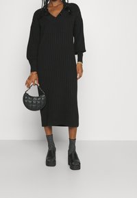 Robe noire en maille côtelée avec décolleté en V, manches bouffantes et longueur mi-mollet, associée à un sac à main noir matelassé et des chaussettes texturées.
