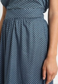 Asymmetrinen mekko, jossa on teksturoitu kangas sinisessä ja mustassa geometrisessä kuviossa, rypytetty vyötärö ja laskeutuva hame.