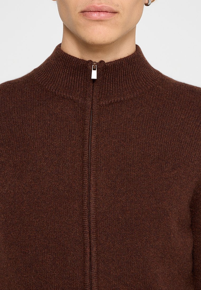 Brauner Reißverschluss-Pullover mit hohem Kragen und gerippter Textur, ausgestattet mit einem silbernen Reißverschluss vorne. Weiches, gestricktes Material.
