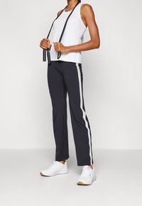 Bílý atletický tílko v kombinaci s černými sportovními kalhotami s bílými postranními pruhy, doplněné bílými teniskami. Hladká látka a pohodlný střih.