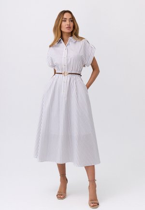 Kobieta stojąca z rękami w kieszeniach, ubrana w białą, pionowo w paski, zapinaną midi sukienkę z krótkimi rękawami i brązowym paskiem, beżowe szpilki.