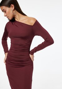 Frau trägt ein weinrotes, gerafftes, einseitiges Langarmkleid mit großen Ohrringen, schaut nach unten und posiert mit den Händen in der Hüfte.