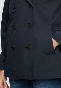 Veste double boutonnage bleu marine avec des boutons noirs, poche latérale et texture lisse. Les poignets présentent un détail de fermeture à boutons.