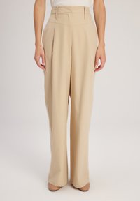 Beige wijde broek met hoge taille, met scherpe plooien en een gladde textuur. Gestyled met een lichte top en nude hakken.