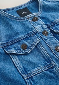 Kék farmerdzseki négy fekete gombbal, két mellrészen zsebfedővel, valamint a gallér belsejében "NEXT" felirattal.