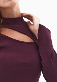 Prążkowany, dopasowany top w głębokim purpurze z wysokim kołnierzem i dekoltem z wycięciem. Długie rękawy z rozszerzającymi się mankietami.