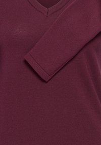 Gros plan d’un pull col en V bordeaux avec une manche longue repliée sur la poitrine montrant une fine texture tricotée.