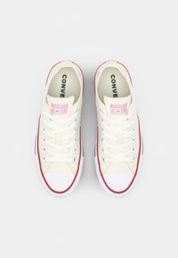 Beige canvas sneakers with white rubber toe caps, red accents, and white laces. Features branding on the tongue and a flat sole.