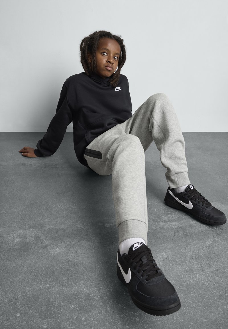 Czarna bluza Nike, szare spodnie dresowe z boczną kieszenią i czarne sneakersy Nike. Gładka tekstura i luźny krój, siedzący na szarym tle.