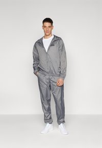 Gray windbreaker with zip, elastic cuffs, and chest logo, paired with matching gray pants, worn with white sneakers. Smooth, lightweight material.