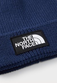Mørkeblå ribbeblåst stoff med et svart rektangulært merke med hvit "THE NORTH FACE" tekst og halvkule-logo.
