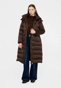 Lång brun dunjacka med pälskantad huva, dragkedja framtill och sidofickor, kombinerad med en mörk stickad tröja och vita vida jeans.