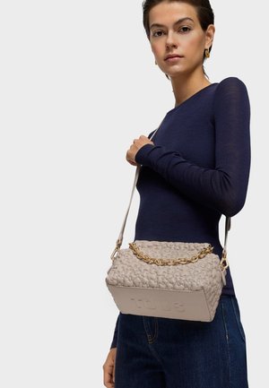 Kobieta w granatowej bluzce z długim rękawem i dżinsach, trzymająca beżową pikowaną torebkę na łańcuszku z złotym paskiem na ramieniu.