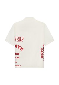 Camicia a maniche corte di colore crema con collo. Presenta una grafica e un testo rosso audace stampati sul retro e sul lato, design minimalista.