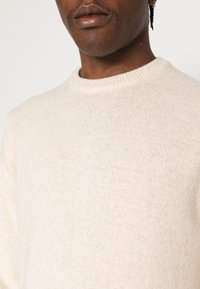 Beige strikket genser med rund hals og ribbet krage. Myk tekstur med små variasjoner i nyanse. Nærbilde av overkroppen.