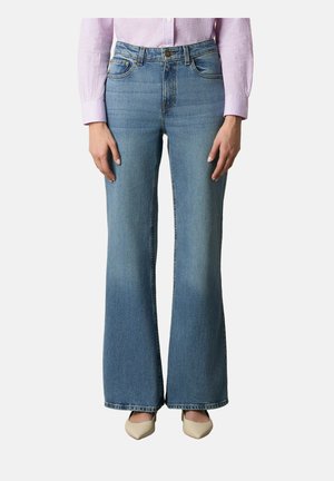 Frau trägt hochtaillierte, hellblaue ausgestellte Jeans mit Vordertaschen, kombiniert mit beigefarbenen spitzen Schuhen und einem rosa karierte Hemd.