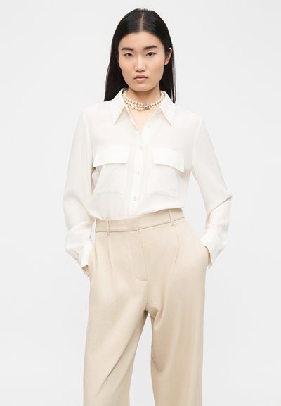 Γυναίκα που φοράει λευκό πουκάμισο με κουμπιά και τσέπες στο στήθος, μπεζ παντελόνι ψηλόμεσο και πολύσειρο κολιέ με μαργαριτάρια, στέκεται με τα χέρια στις τσέπες.