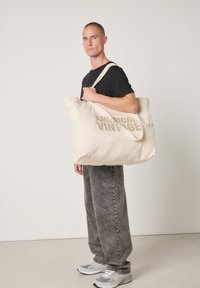 Sac fourre-tout en toile beige avec le texte "AMERICAN VINTAGE" imprimé en marron clair, grande taille, texture douce, et double bandoulière.