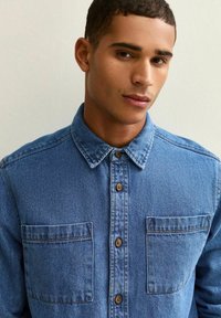 Lichtblauwe denimshirt met knoopsluiting aan de voorkant, twee borstzakken en een klassieke kraag. De stof heeft een gestructureerde afwerking.