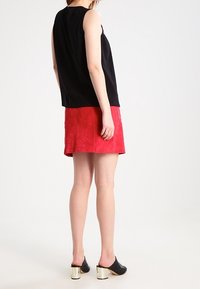 Top senza maniche nero abbinato a una gonna in scamosciato rosso. Il modello indossa mule nere con tacco a blocco metallico. Design minimalista e colori audaci.