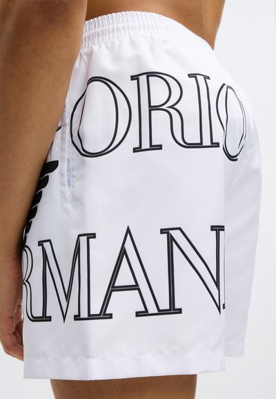 Λευκό μαγιό σορτς με μεγάλο μαύρο κείμενο "Giorgio Armani." Το ύφασμα είναι απαλό, με ελαστική μέση και πλαϊνές τσέπες.