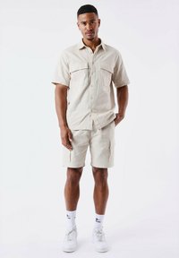 Camisa beige de manga corta con botones y dos bolsillos en el pecho, combinada con pantalones cortos cargo a juego. Usada con zapatillas blancas y calcetas.